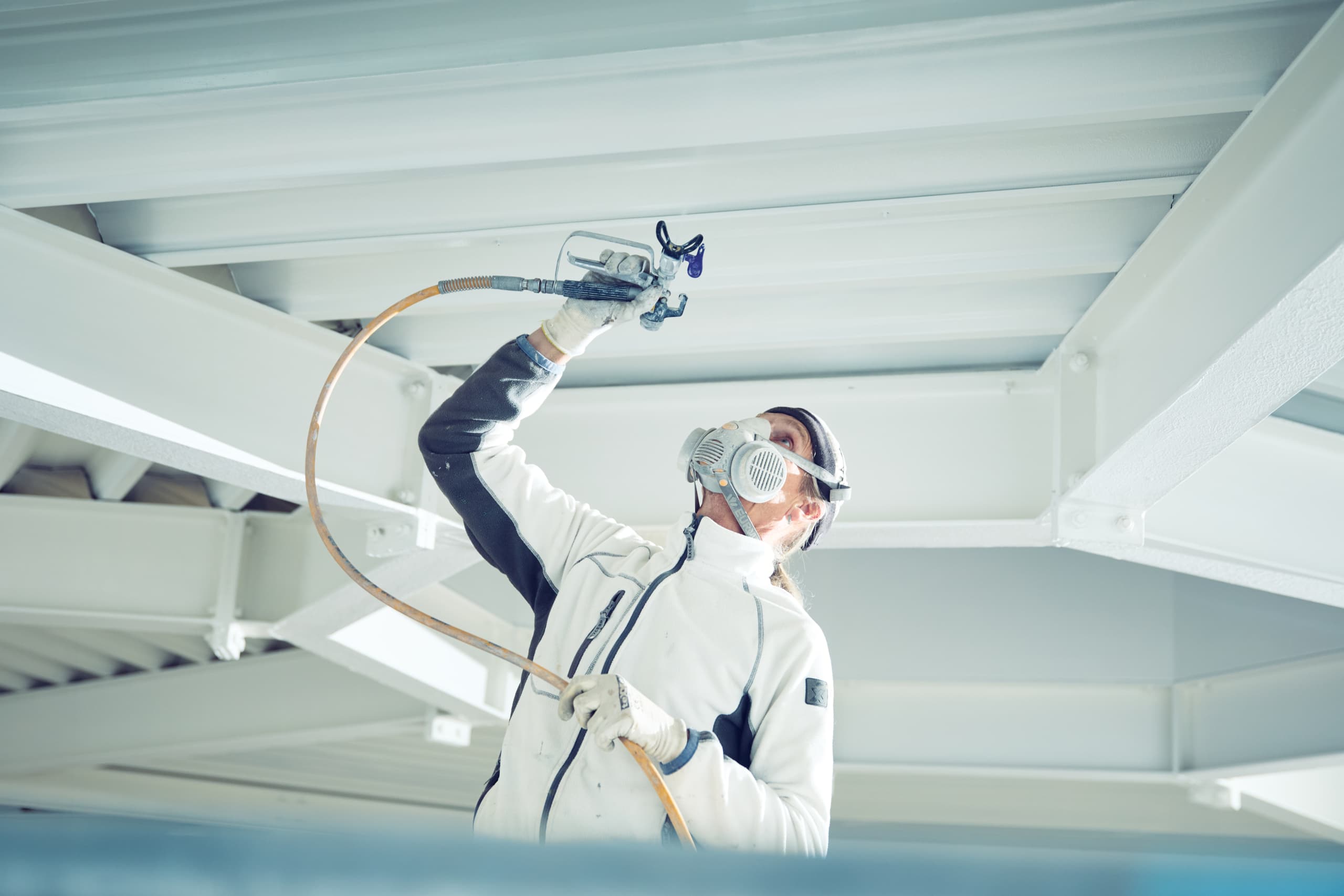 Maler mit Atemschutzmaske besprüht weiße Stahlkonstruktion mit Airless-Spritzgerät