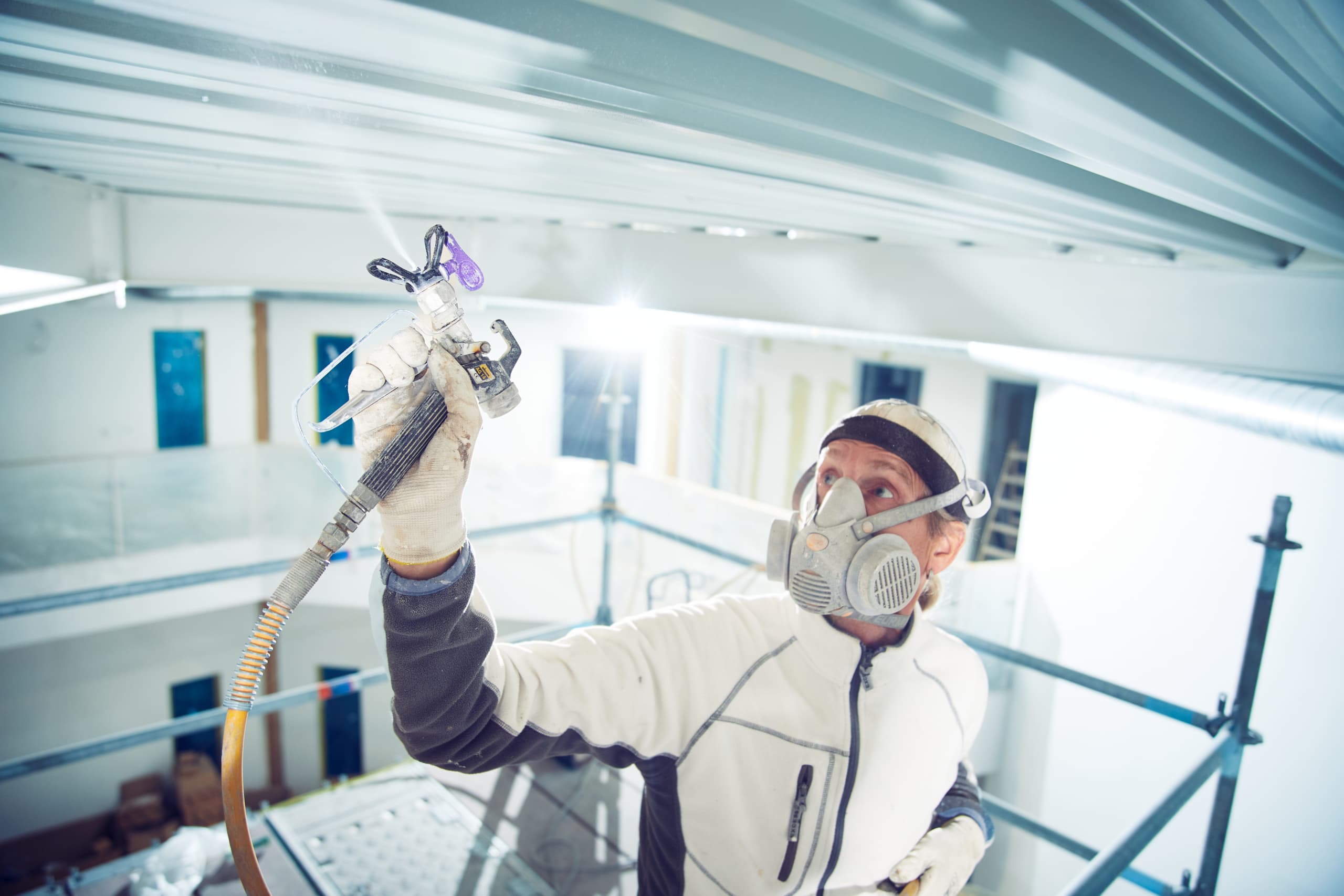 Maler mit Atemschutzmaske sprüht Farbe an eine Decke auf einer Baustelle
