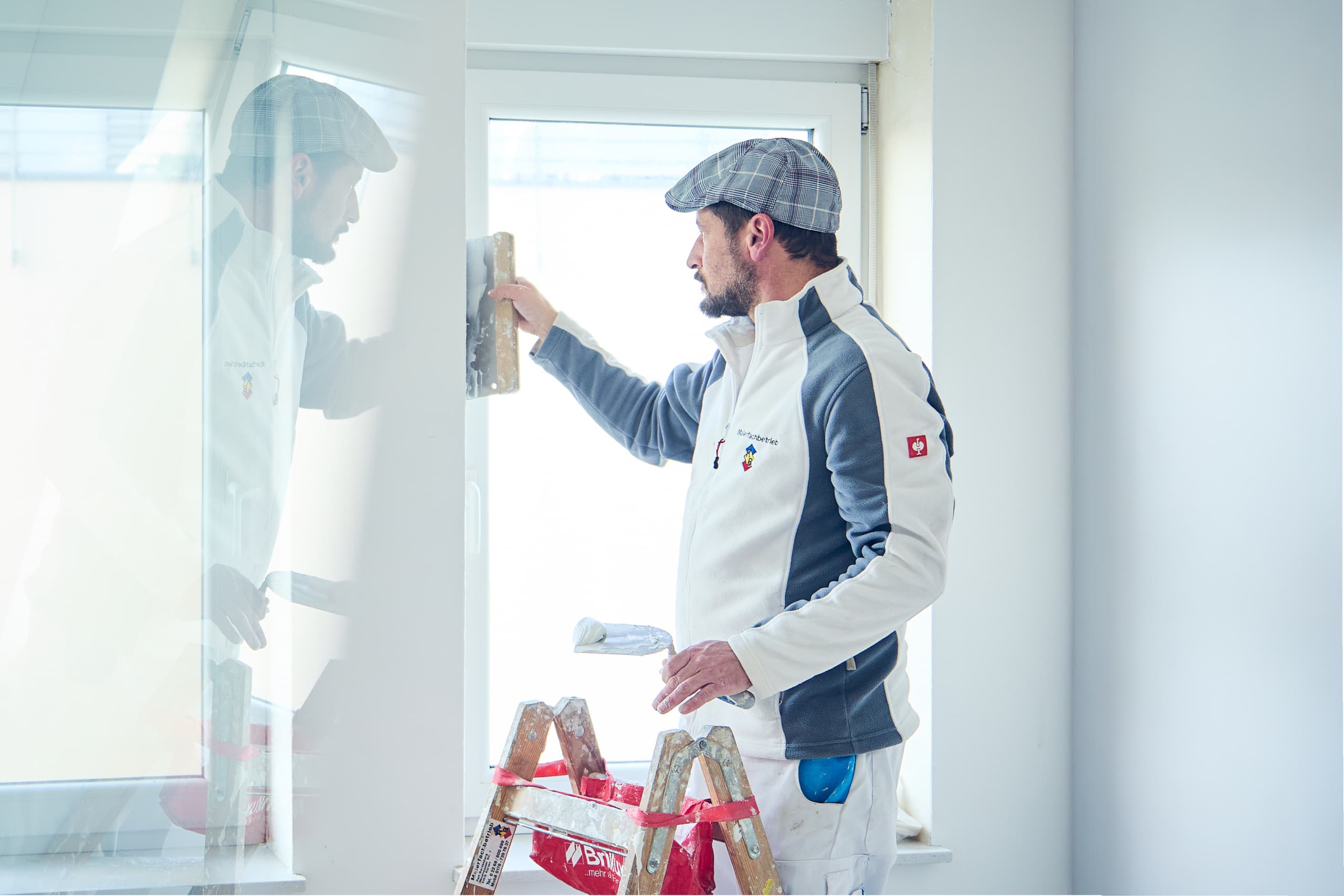 Maler in Arbeitskleidung verspachtelt eine Wand neben einem Fenster