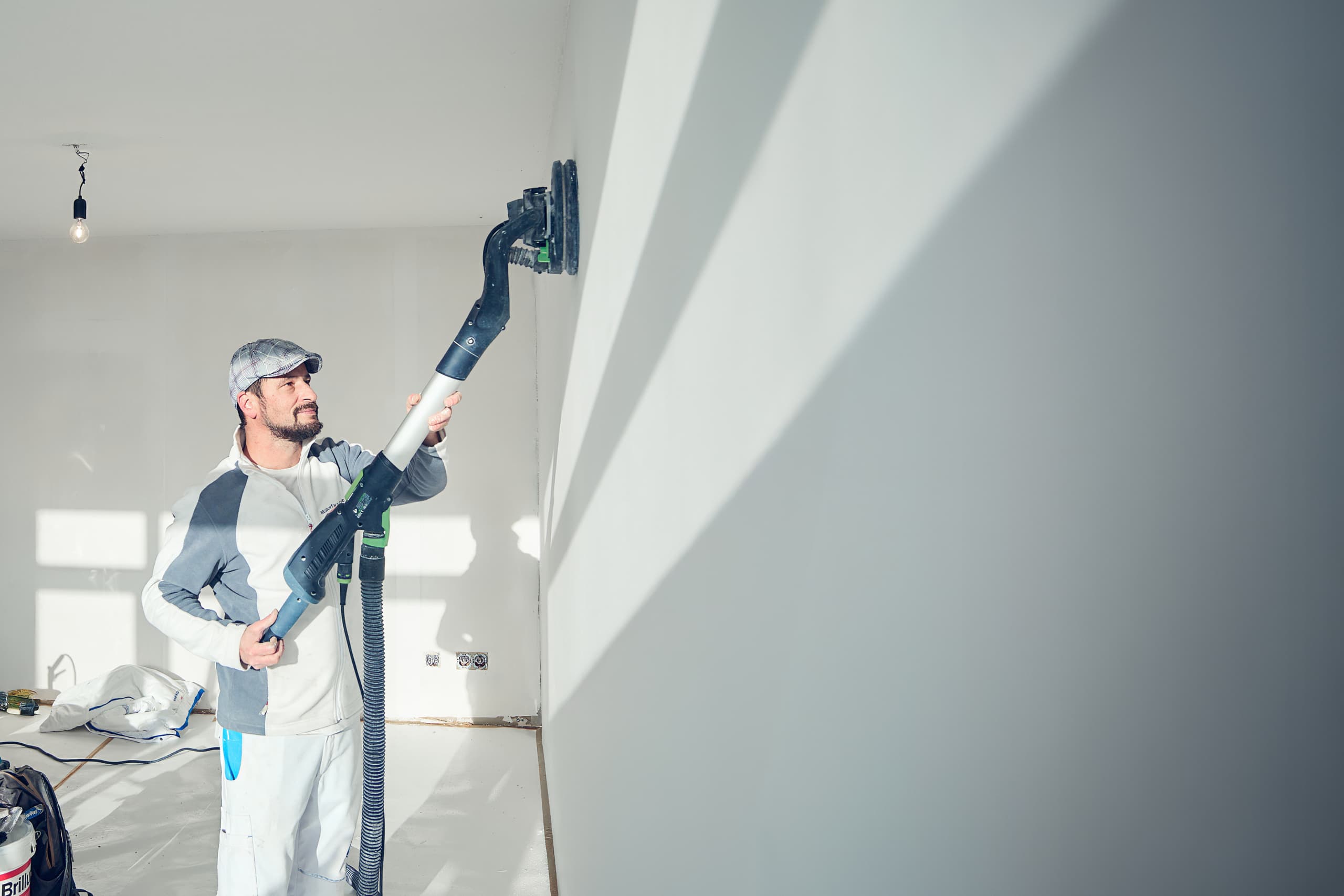 Maler in Arbeitskleidung schleift mit Langhalsschleifer eine Wand in einem hellen Raum