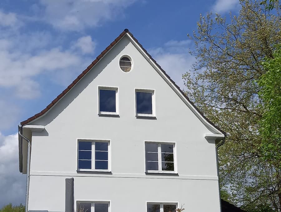 Weiß verputztes Einfamilienhaus mit Spitzgiebel, rotem Ziegeldach und Rundfenster vor blauem Himmel