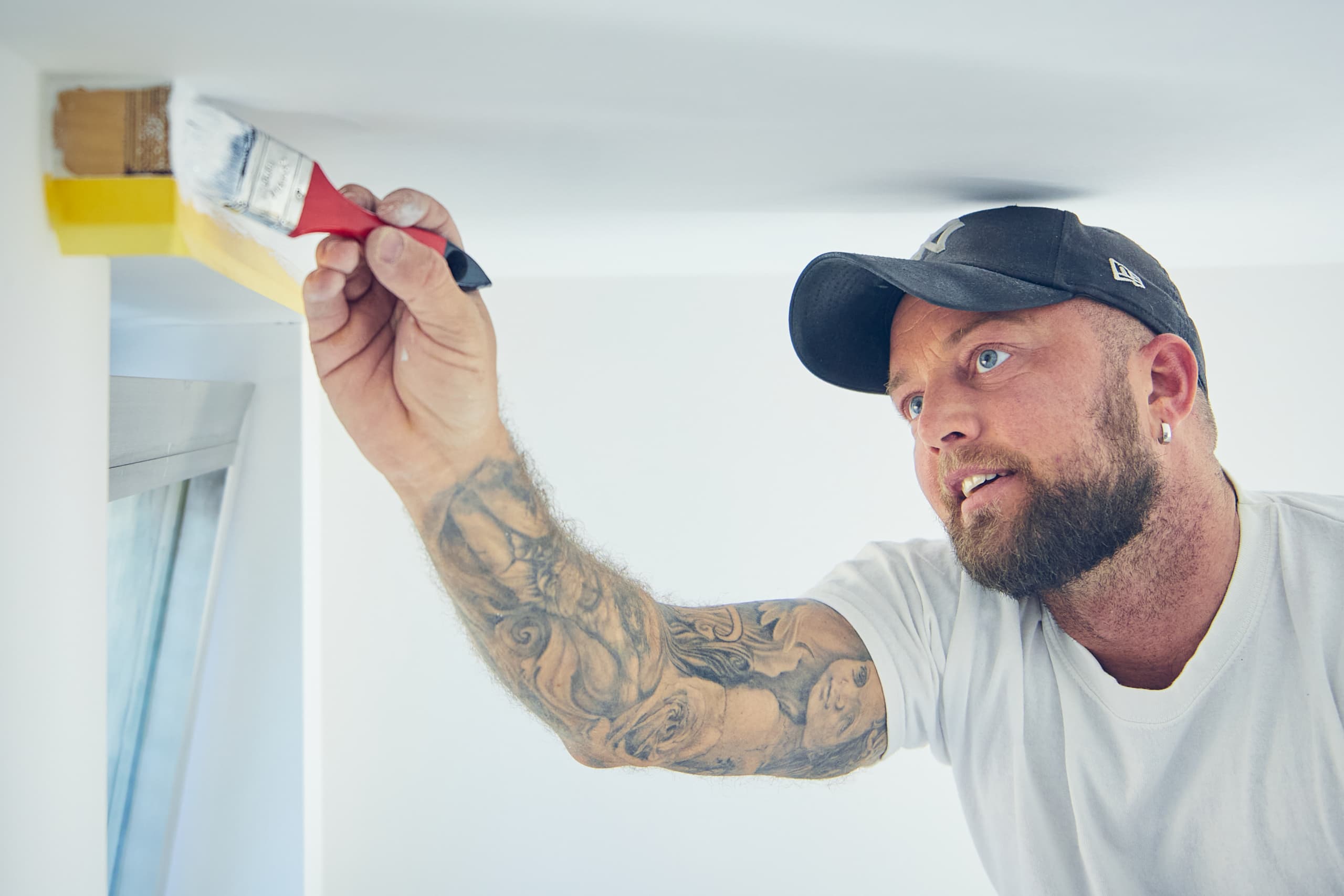 Maler mit Basecap und tätowiertem Arm streicht konzentriert eine Kante an der Decke