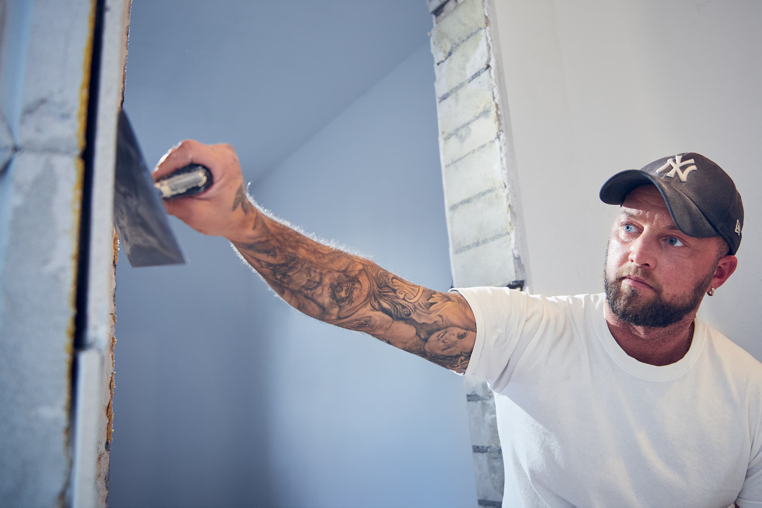 Maler mit tätowiertem Arm und Cap verspachtelt eine Wand mit einer Glättkelle